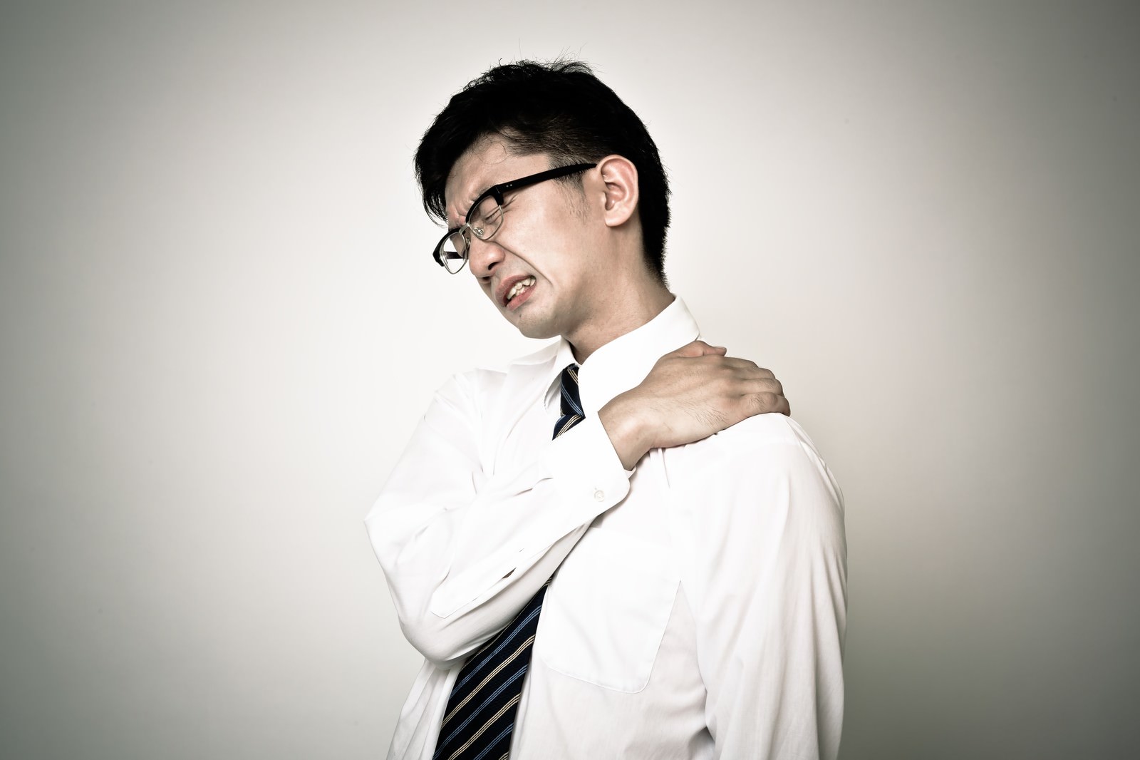 右手で左肩を押さえて顔をしかめる肩こりのスーツ男性［モデル：OZPA］
