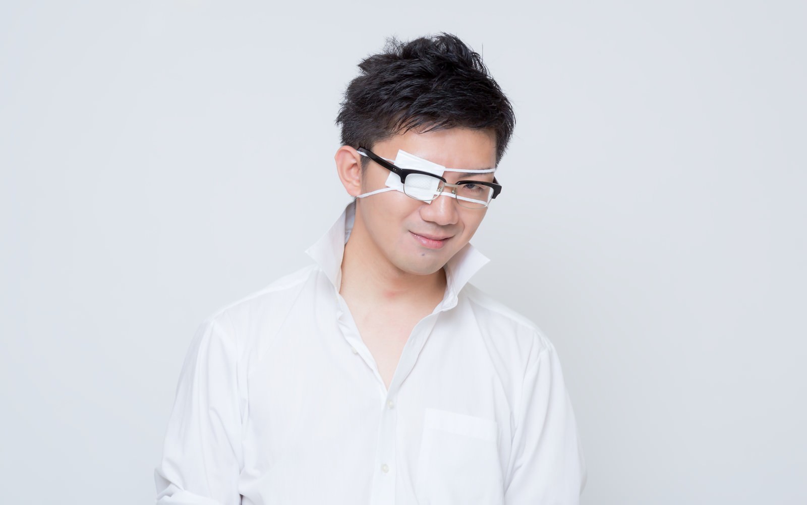 眼鏡の片側レンズに白い眼帯を貼り、薄ら笑いで斜めに視線を向ける白シャツの男性［モデル：OZPA］