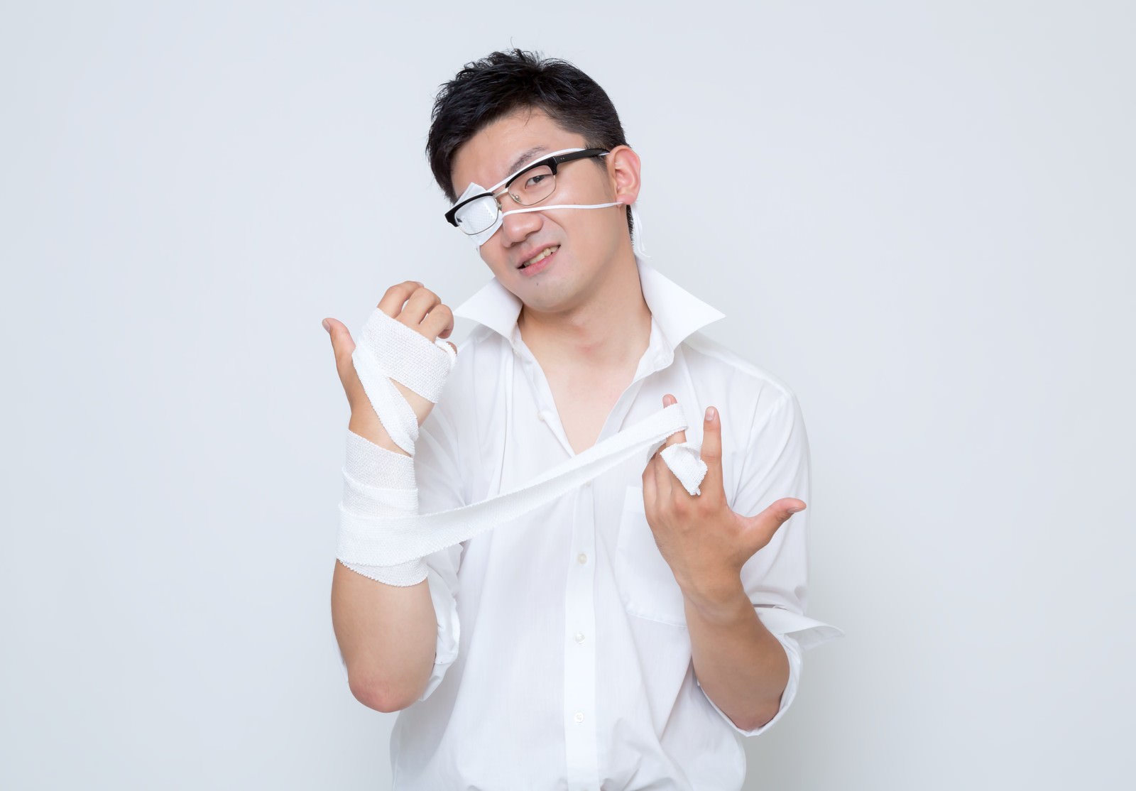 眼鏡をかけた白いワイシャツの男性が、右腕の白い包帯を両手で解いている［モデル：OZPA］