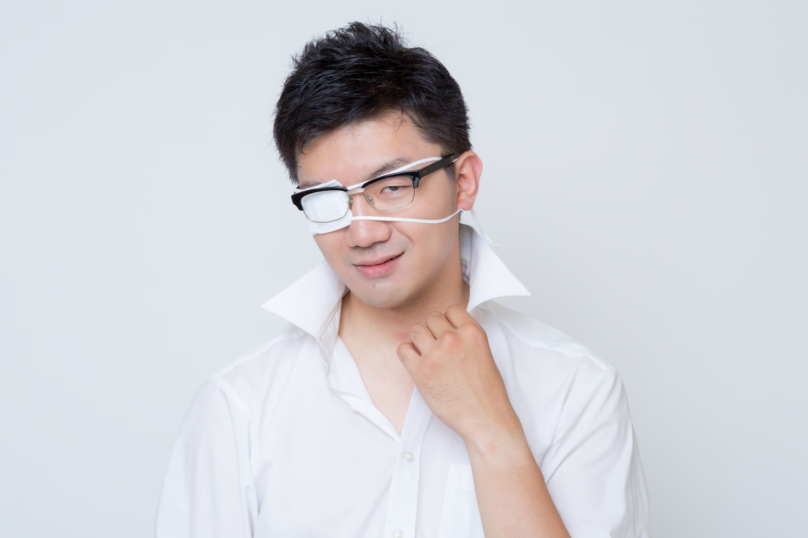 左目に眼帯をつけた眼鏡の男性が首元に手を添えて余裕の笑みを見せる様子［モデル：OZPA］