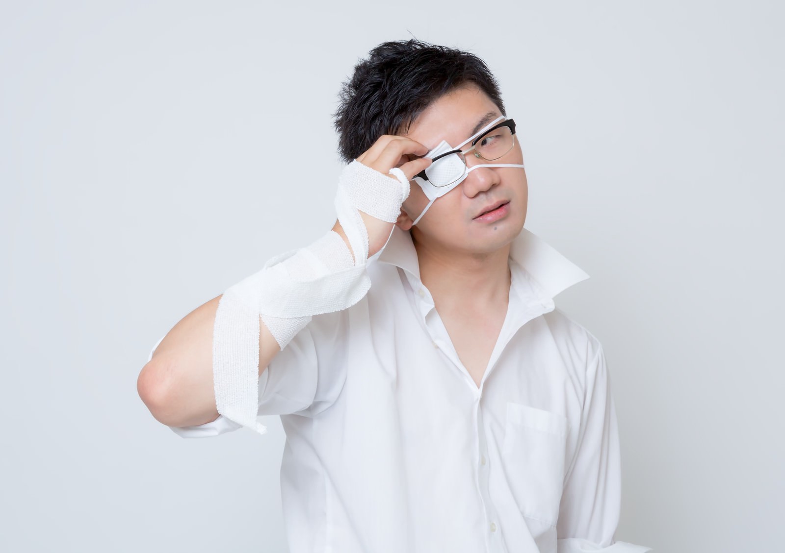 白い眼帯と包帯を着けた男性が、眼鏡をかけながら右目に手を当てて上を見つめている［モデル：OZPA］