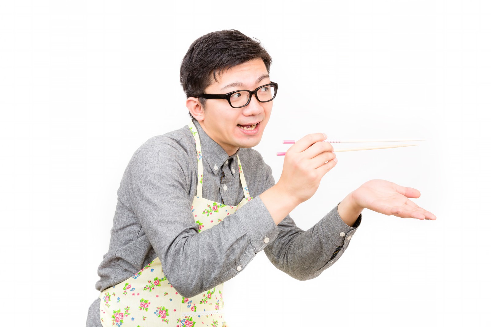 グレーシャツとフラワー柄エプロン姿で、ピンク色の菜箸を手に笑顔で味見をする眼鏡の男性［モデル：OZPA］