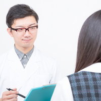 青緑のクリップボードを持ち後ろ姿の女性を診察する医師の写真