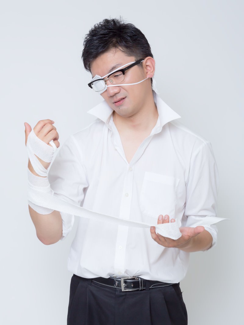 眼帯をつけ、両手首に包帯を巻いた男性が包帯に目を落としながら立つ［モデル：OZPA］