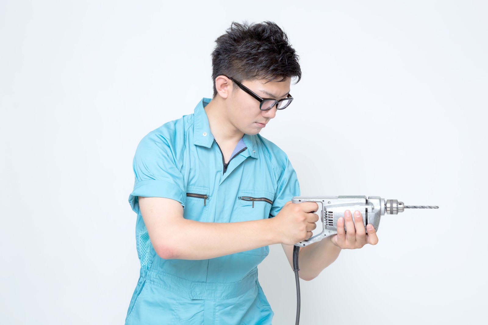 青い作業着の男性が電気ドリルを両手で構えている［モデル：OZPA］