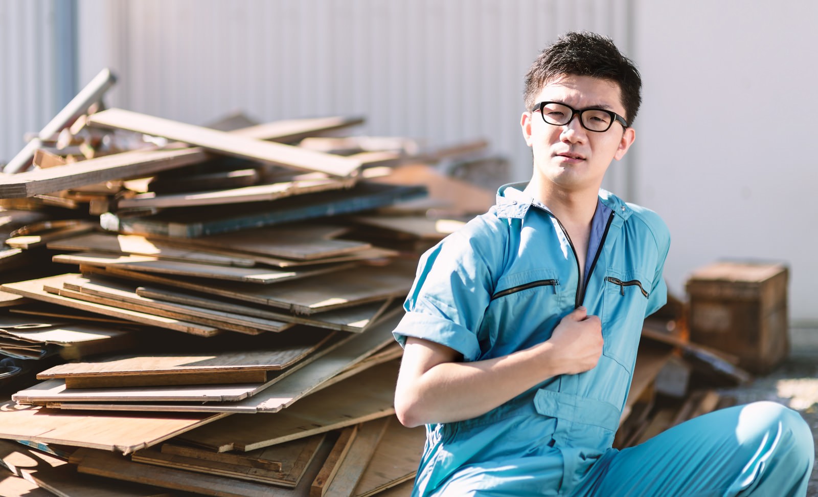 廃材置き場で両腕を組む青い作業着姿の男性［モデル：OZPA］