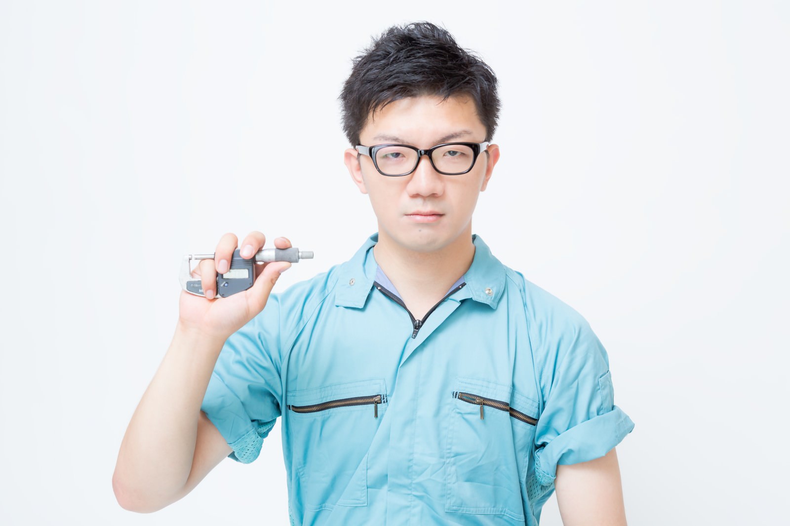 マイクロメーターを持つメガネの男性作業員が水色の作業着姿で立っている［モデル：OZPA］