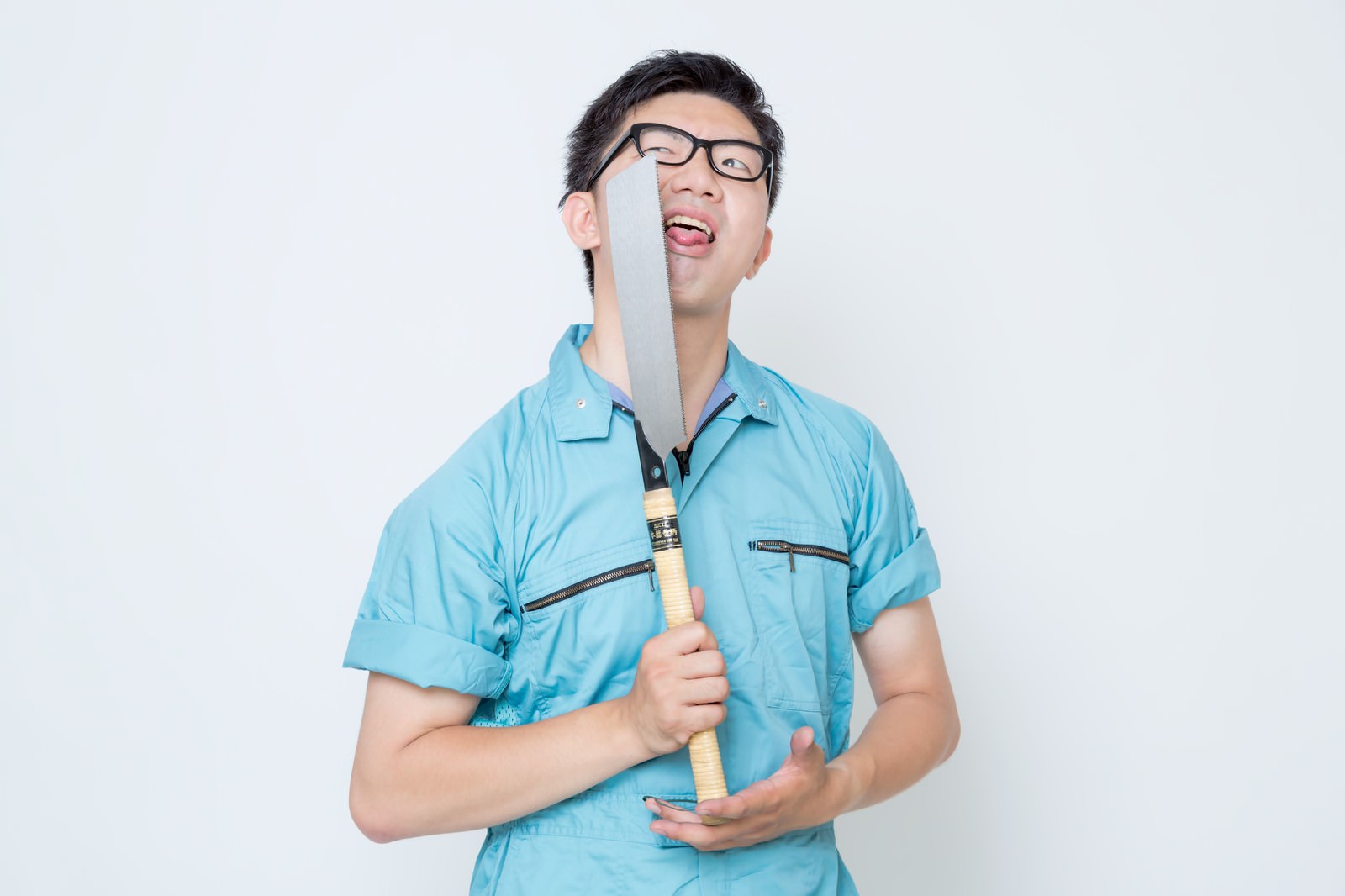 ノコギリを構える青い作業着の男性がメガネをかけ口を開けている［モデル：OZPA］