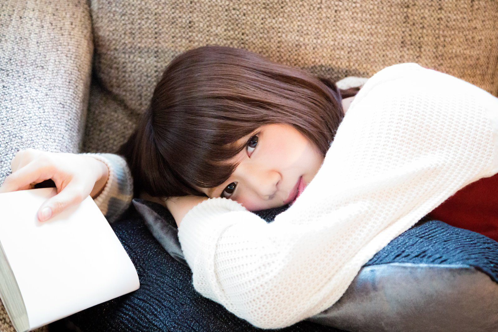 白いニットを着た女性がソファーで本を読みながら片手を伸ばし、相手に呼びかけている［モデル：みき。］