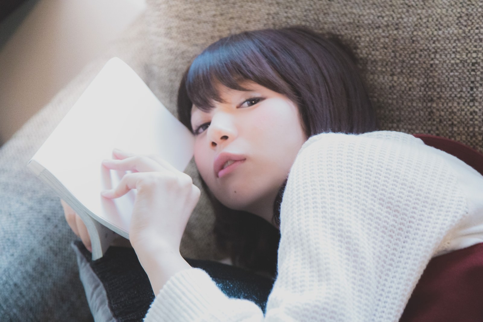 ソファーに横たわるニット姿の女性がマンガを読んでいる［モデル：みき。］
