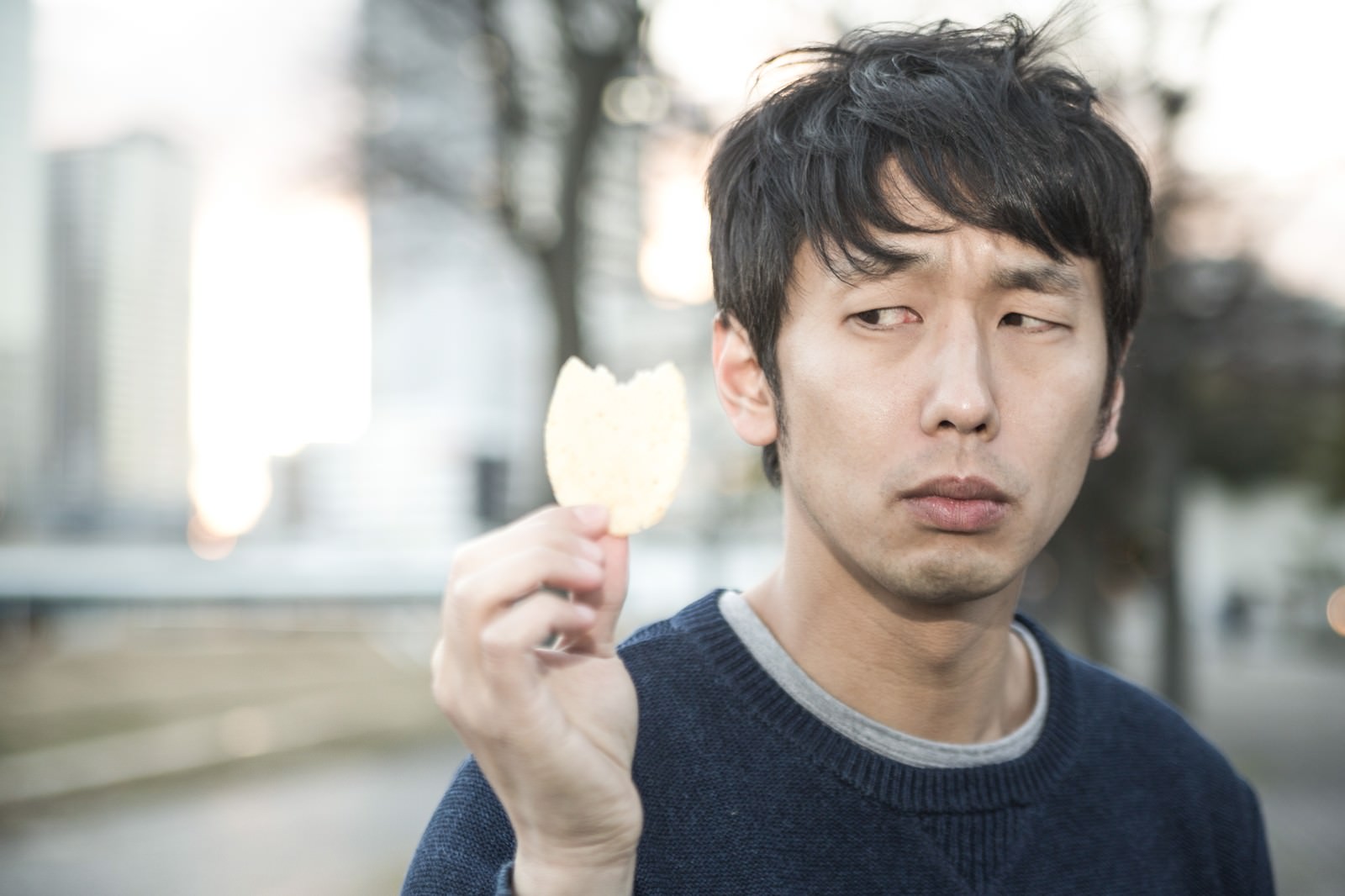 紺色ニットの男性がポテトチップスを片手に持ち、下を向いて考え込む表情で街で立つ［モデル：大川竜弥］