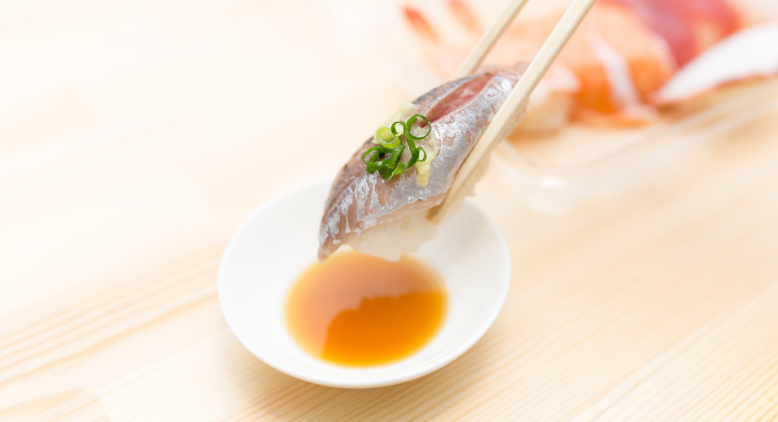 箸でつまんだネギのせアジの切り身が小皿の醤油の上にあり背景に寿司がぼけている