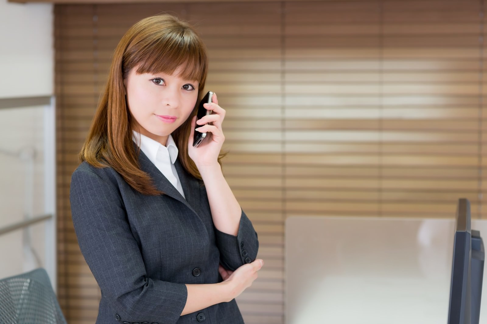 グレーのスーツの女性がオフィスで黒い携帯電話を右手で耳に当てている［モデル：暢子］