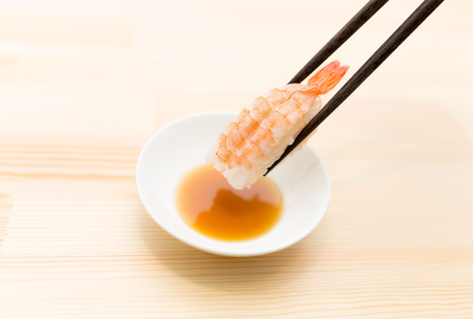黒い箸でつまんだエビの握り寿司を小皿の醤油につけようとしている