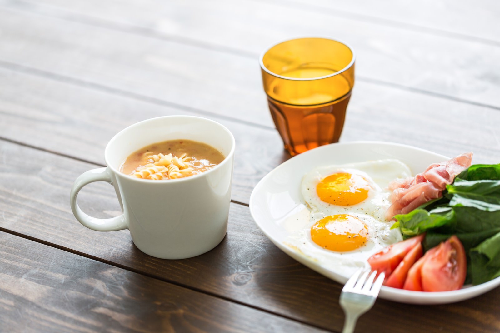 白いプレートに盛られた目玉焼きとハム、野菜、背景にスープとドリンク