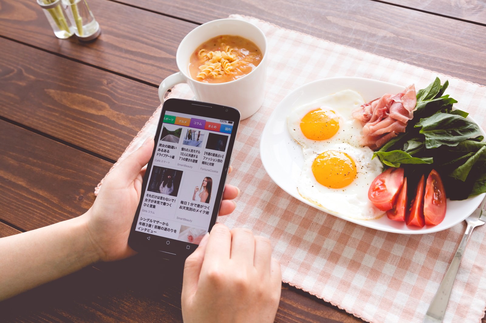 朝食時にスマートフォンでスマニューアプリを開く女性の手。テーブルに並ぶスープと目玉焼き