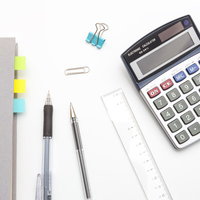 白い机に並べた電卓とノートと文房具の俯瞰の写真