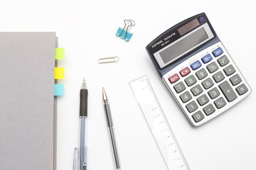 白い机に並べた電卓とノートと文房具の俯瞰