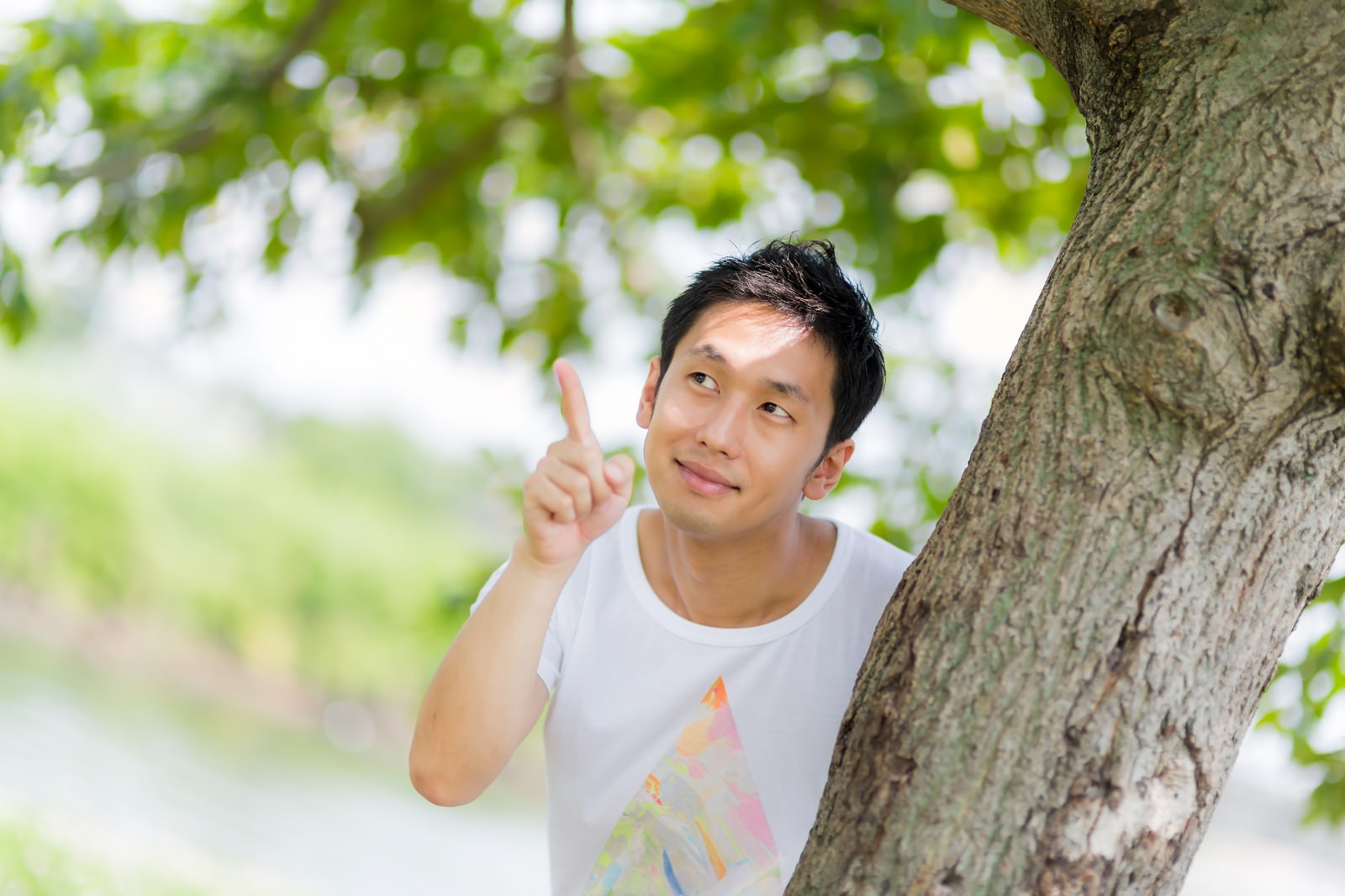 木の根元から身を乗り出して人差し指を立てて上を向く男性［モデル：大川竜弥］