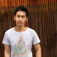茶色い壁の前に立つ白Tシャツの男性の写真