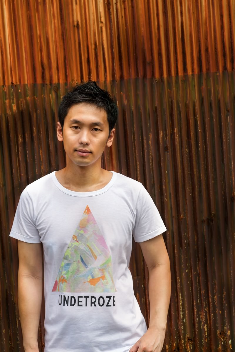 廃工場の前でファッションブランドのTシャツを着た男性の正面顔写真［モデル：大川竜弥］