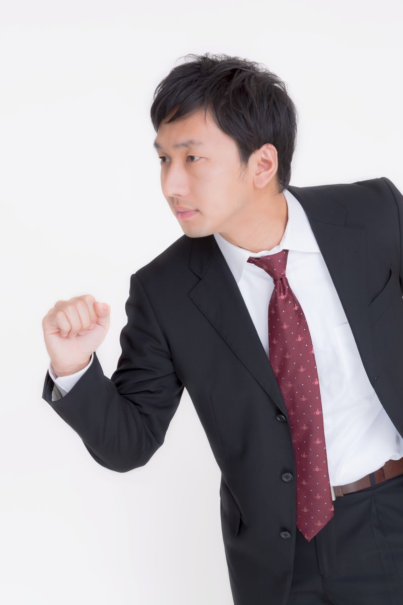 黒いスーツに赤いドット柄ネクタイの男性が横向きで左手の拳を前に出している［モデル：大川竜弥］