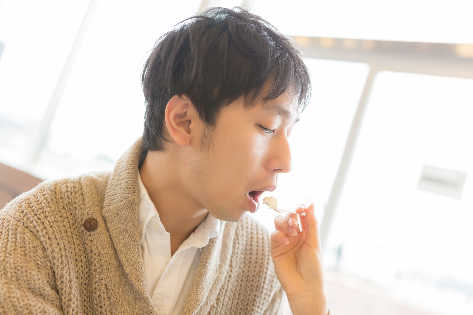 カフェの窓辺でベージュのカーディガンの男性が横向きに座り手を口元に当てている［モデル：大川竜弥］