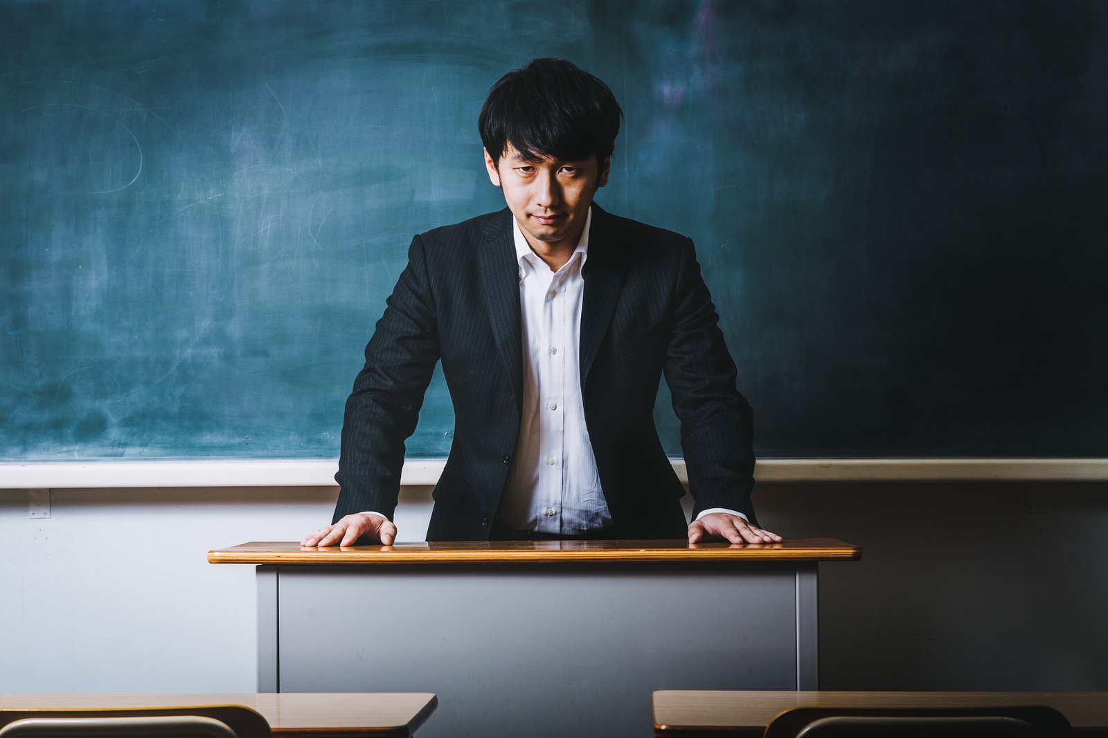 黒板の前で微笑む高校教師の男性。黒いスーツと白いシャツを着て教卓に両手をついている［モデル：大川竜弥］