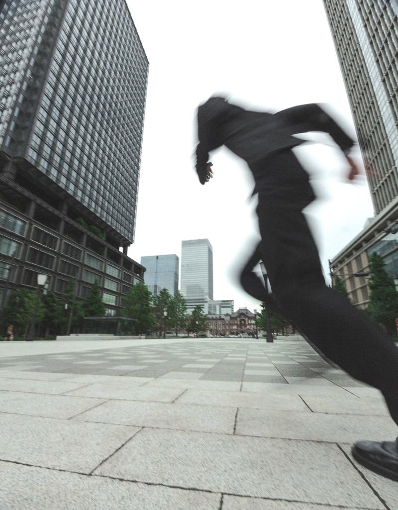 高層ビルに囲まれた広場を黒いスーツ姿で全力疾走するビジネスマン