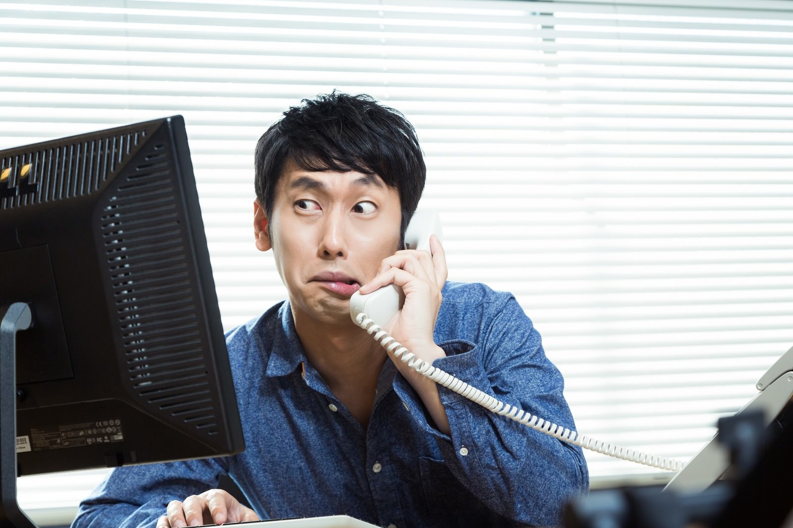 デスクで受話器を耳に当てキーボードを操作する青シャツのエンジニア［モデル：大川竜弥］