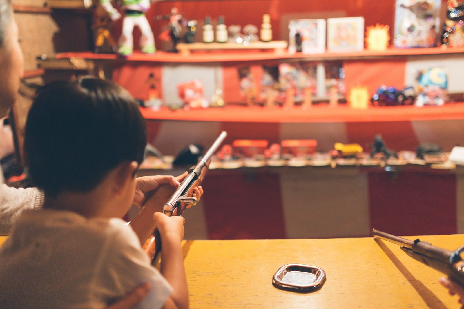 お祭りの赤い出店で銃を構える黒髪の子供。黄色いテーブルと景品が並ぶ棚が背景