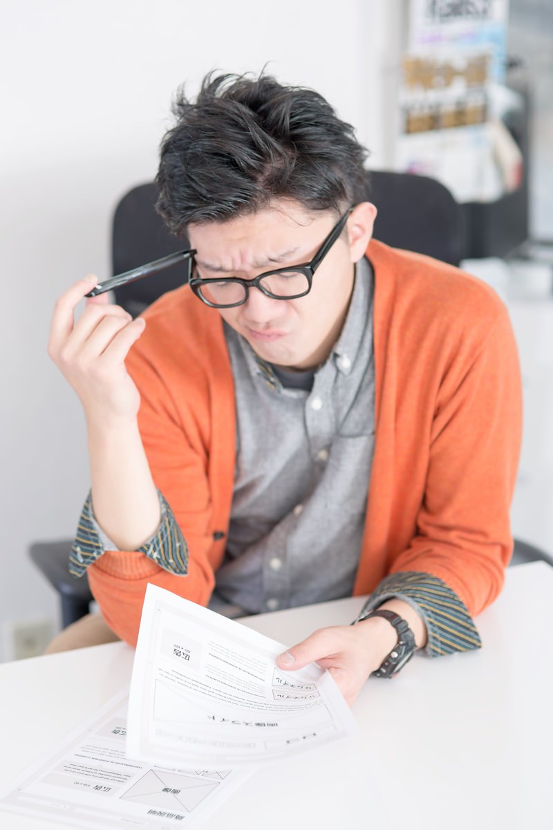 机の前で眼鏡を持ち上げ書類を見るオレンジカーディガンの男性［モデル：OZPA］