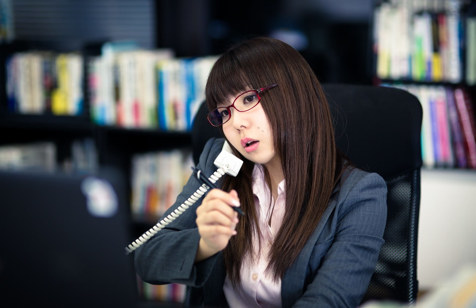 眼鏡をかけた女性がグレーのスーツ姿で本棚の前で黒い電話の受話器を持っている［モデル：Lala］