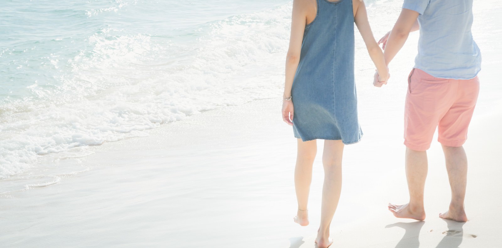波打ち際で手をつなぐカップル。女性は青いワンピース、男性はピンクのショーツを着用