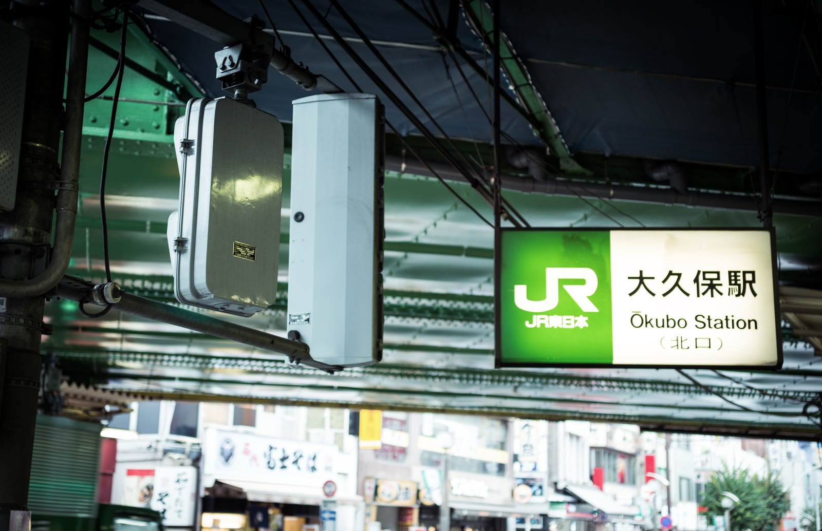 夜間に緑色に照らされたJR大久保駅前の駅名表示板と白い配電盤
