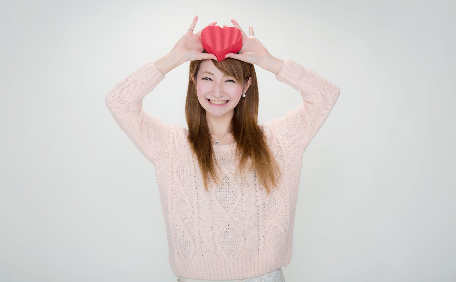 ピンク色ニットの女性がハート型の赤いプレゼントボックスを頭上に掲げ、笑顔を向けている［モデル：to-ko］