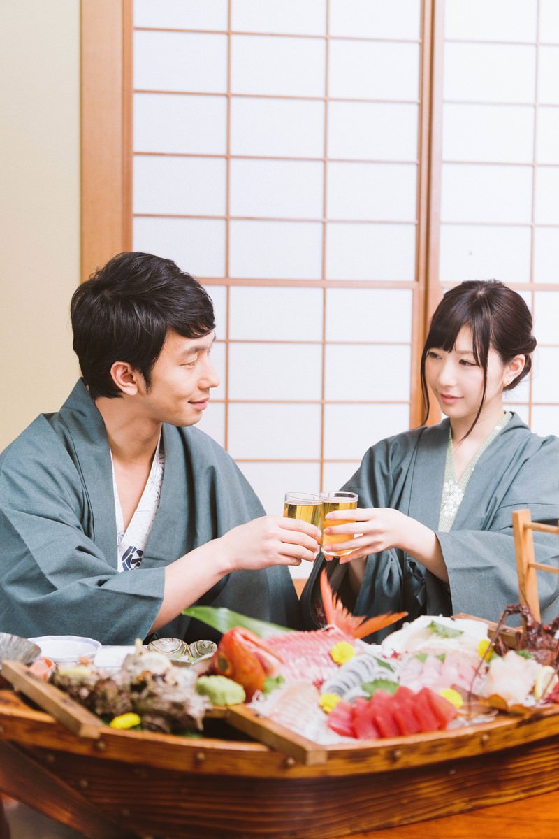 浴衣姿のカップルが旅館の宴会で乾杯をしている。テーブルに舟盛りの海鮮料理が並んでいる［モデル：大川竜弥 モデルリリース］