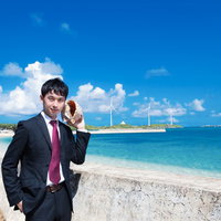 白い砂浜と風車が見える海辺で貝殻を耳に当てるスーツの男性の写真