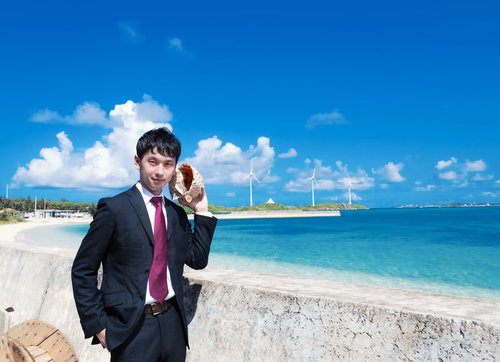 白い砂浜と風車が見える海辺で貝殻を耳に当てるスーツの男性