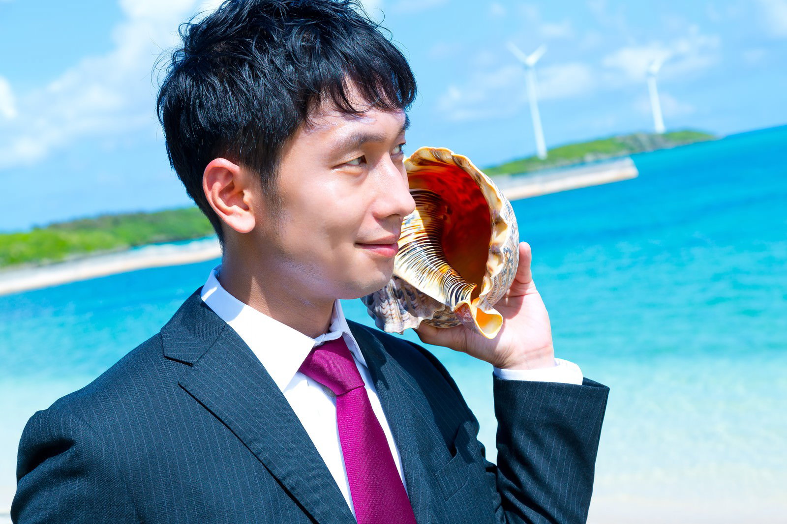 海岸で貝殻を耳に当てるスーツ姿のビジネスマン［モデル：大川竜弥］