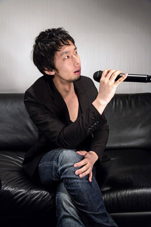 ソファに座りマイクを持つ黒ジャケットの男性