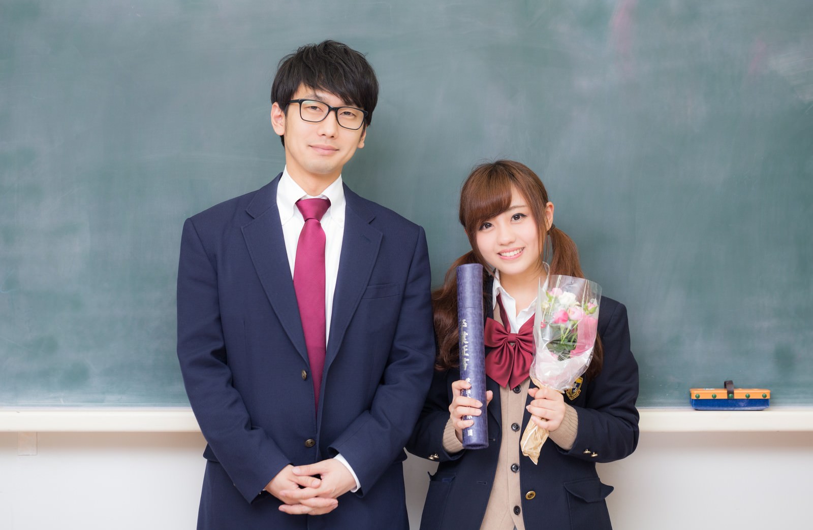 教室の黒板前に立つ制服姿の高校生2名。左は眼鏡と赤紫ネクタイの男性、右は花束と卒業証書を持つ女性［モデル：大川竜弥 河村友歌］
