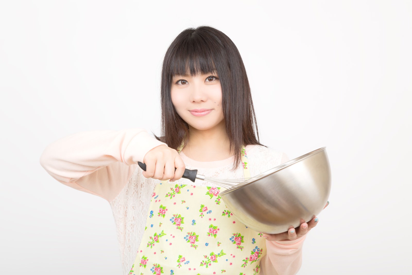 花柄エプロンの女性が銀色のボウルと泡立て器を持って笑顔を見せている［モデル：Lala］