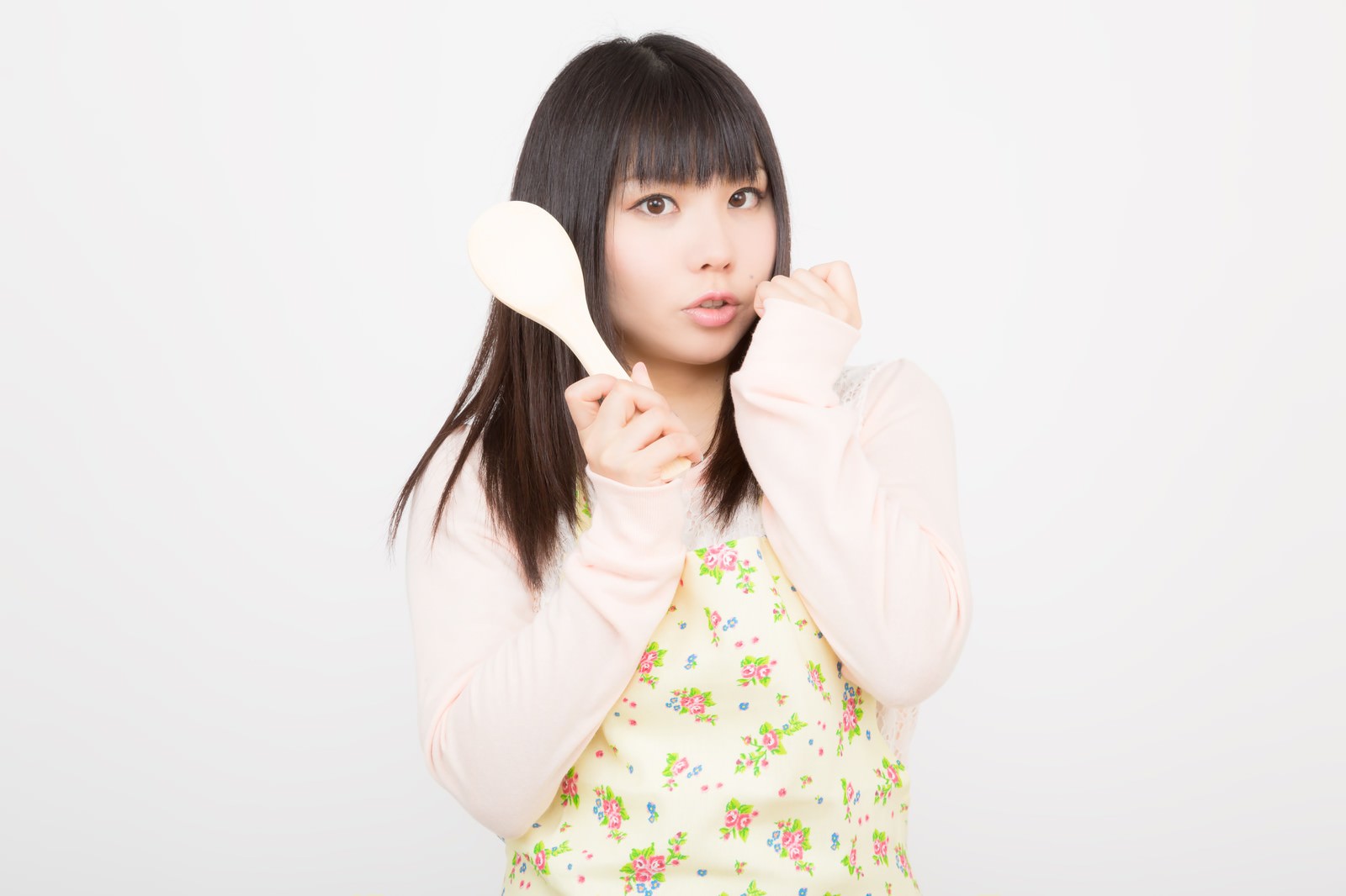 黄色い花柄エプロンの女性が白いしゃもじを頬に当てて正面を向いている［モデル：Lala］