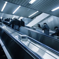 黒やグレーの服の人々が手すりに手を置いて上るエスカレーターの写真