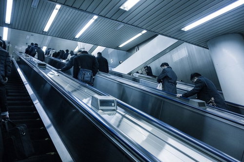 黒やグレーの服の人々が手すりに手を置いて上るエスカレーター