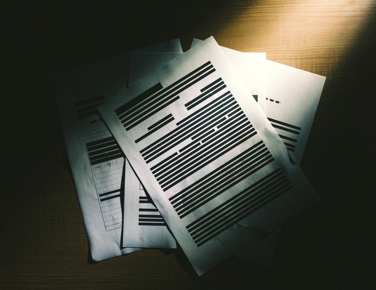 黒塗りされた機密情報が含まれた複数の書類が木製の机の上に積み重ねられている