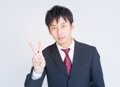 白背景で右手ピースサインをするスーツのビジネスマン