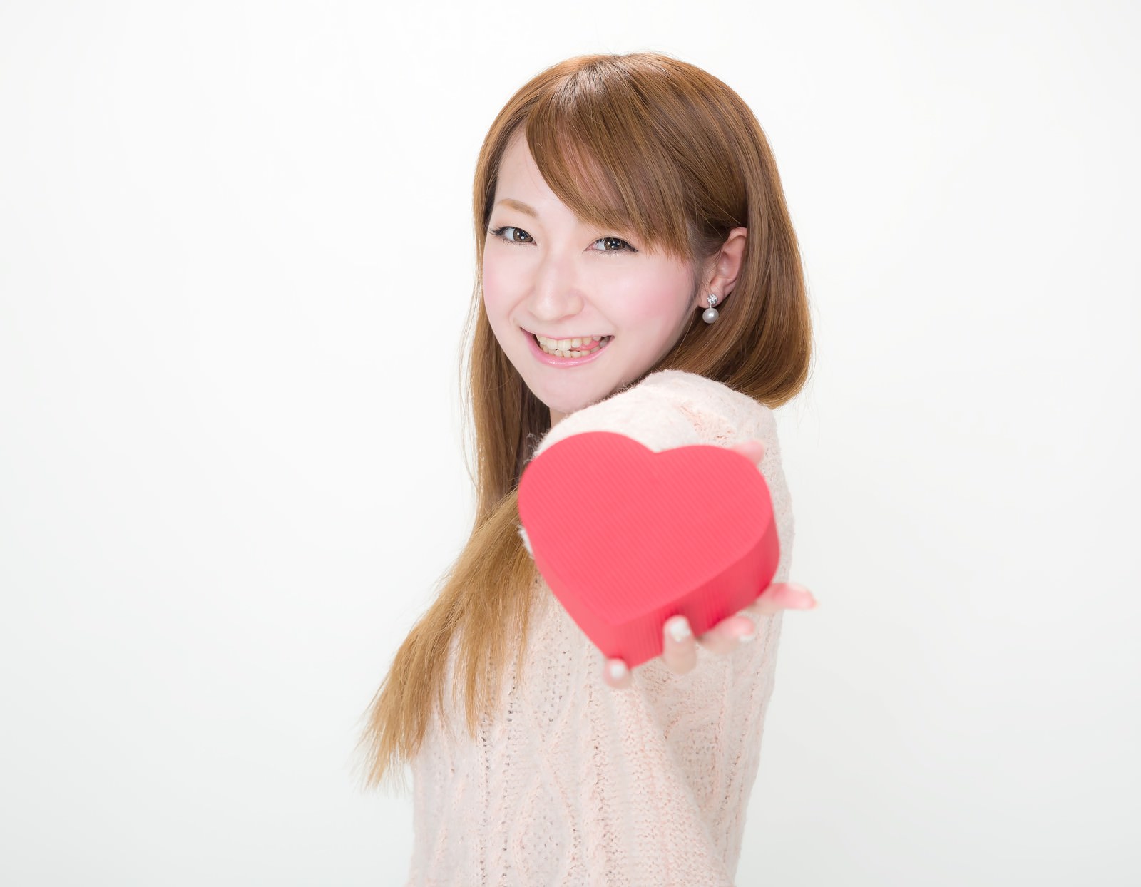 ベージュのニットを着た女性が赤いハート型チョコを手に笑顔で振り返る［モデル：to-ko］