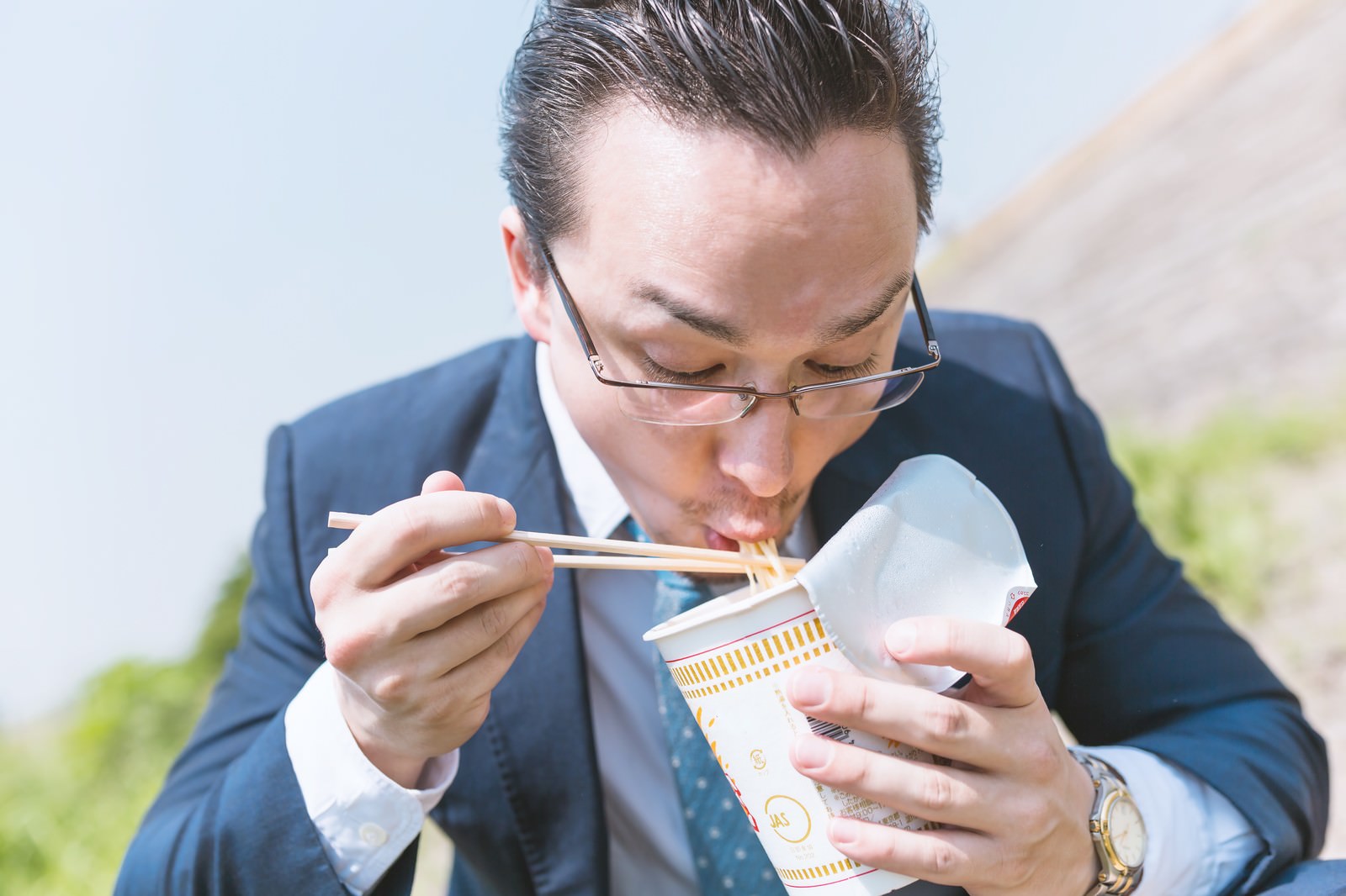 屋外でカップラーメンをすするスーツの男性［モデル：Max_Ezaki］